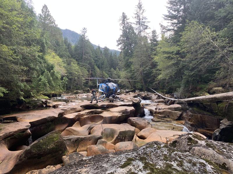 Blue helicopter on a rocky riverbed surrounded by forest.