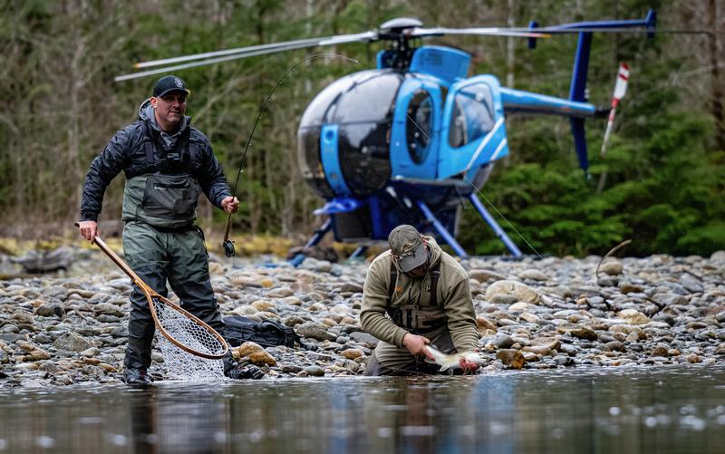 Angler netting a fish on a riverbank near a blue helicopter.