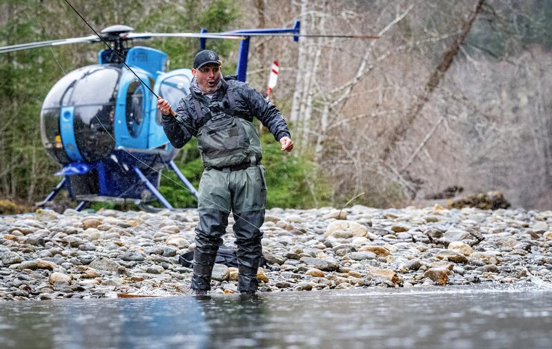 Angler in waders fishing in a river with a helicopter parked on the shore nearby.