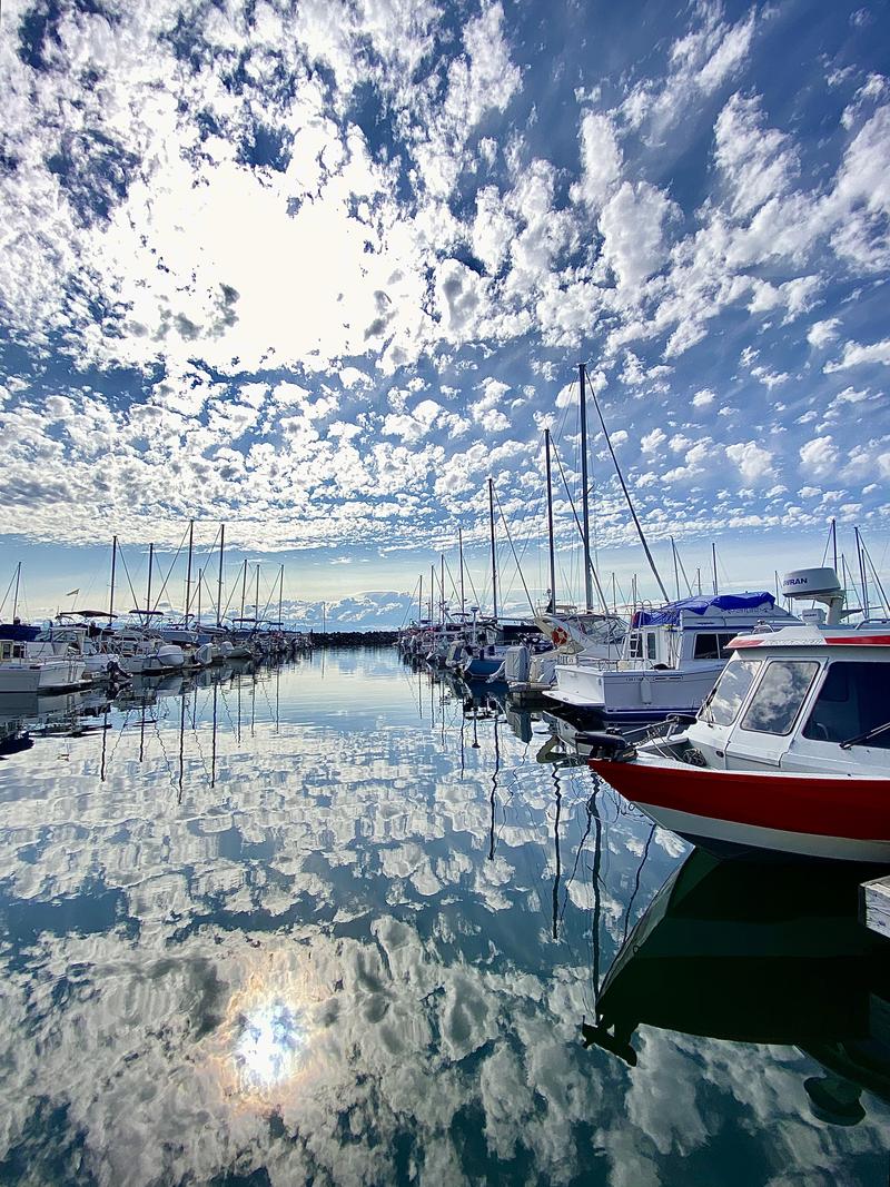 Marina with boats docked in calm water reflecting a blue sky with scattered clouds.