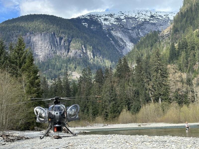 Blue helicopter parked on a gravel bar with mountains in the background.