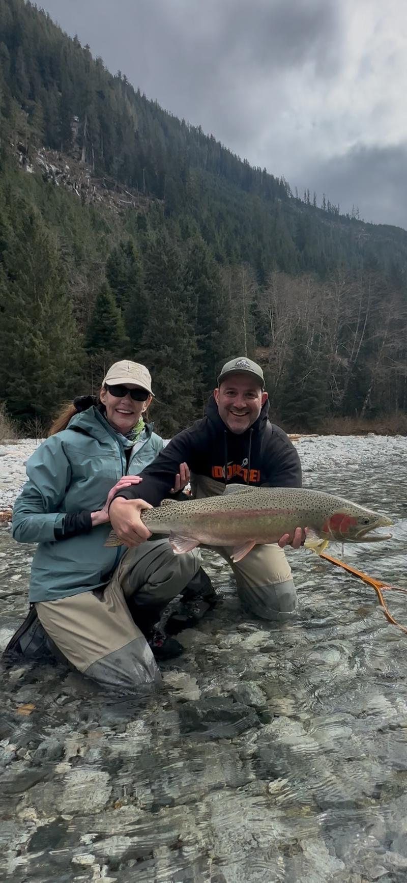 Couple kneeling in a river holding a salmon with a forest backdrop.