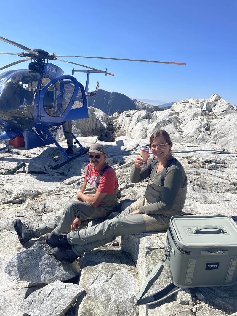 Two people relaxing on rocks near a blue helicopter with drinks.