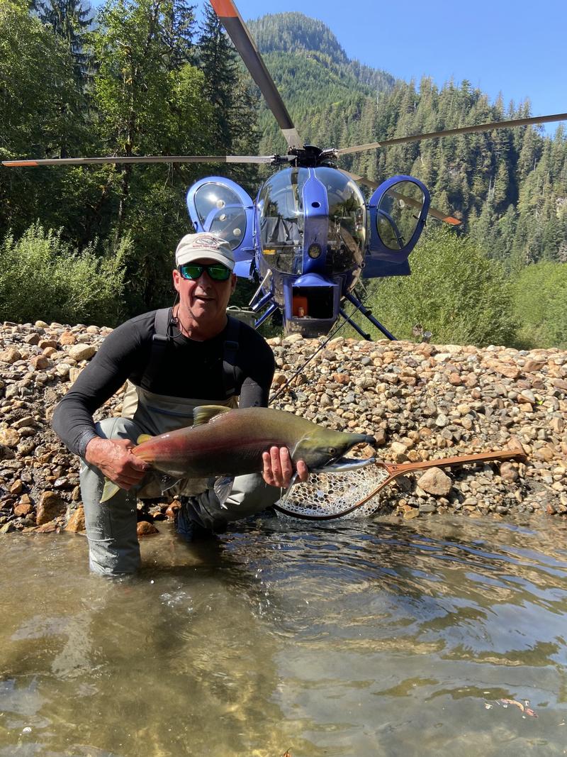 Angler holding a fish in a river with a blue helicopter in the background.