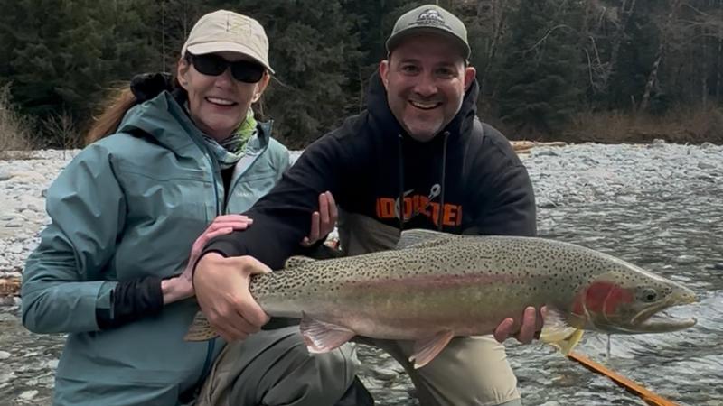 Man standing in a river holding a large fish he caught with a helicopter in the background