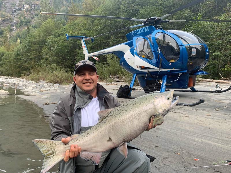 Fisherman holding a large salmon on a beach with a blue helicopter in the background.