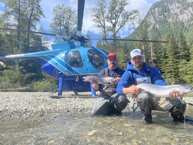 Two anglers posing with a large fish in front of a blue helicopter.